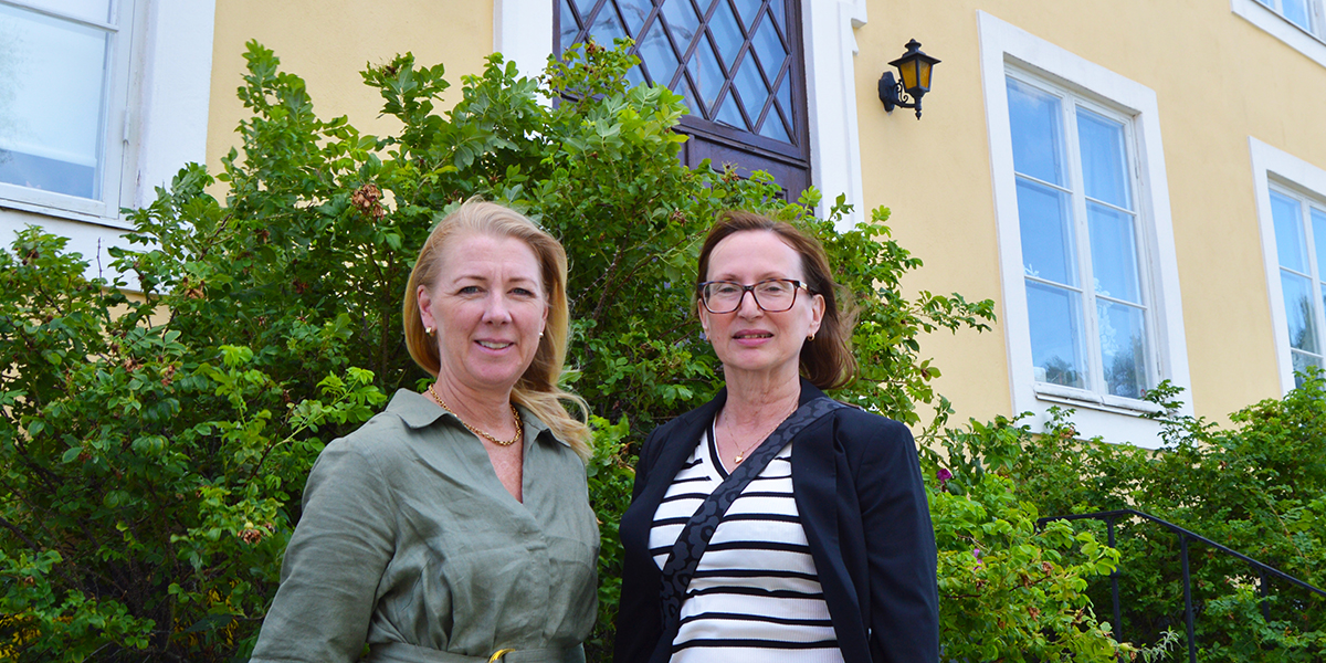 Två kvinnor står bredvid varandra och tittar mot kameran, framför en grön buske. De är fotograferade utomhus vid en gul byggnad med vita fönster, en ytterlampa och en dörr i bakgrunden.
