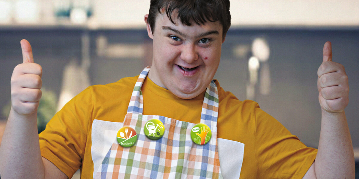 Leende person i kök lyfter båda tummarna upp och bär rutigt förkläde över gul tröja. På bröstet sitter knappar med texten “MatGlad 2.0” och “TAKK”, samt en grön symbolknapp.