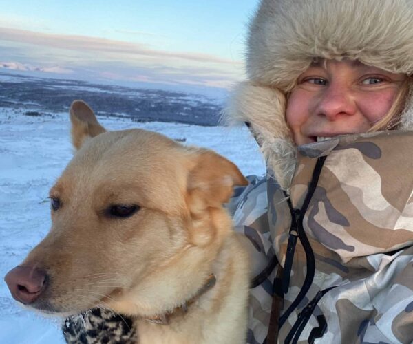 Leende person i kamouflagejacka med stor pälsluva tar en selfie och håller en ljusbrun hund nära sig. De står utomhus i vinterkyla, med snötäckta fjäll och blekblå himmel i bakgrunden.
