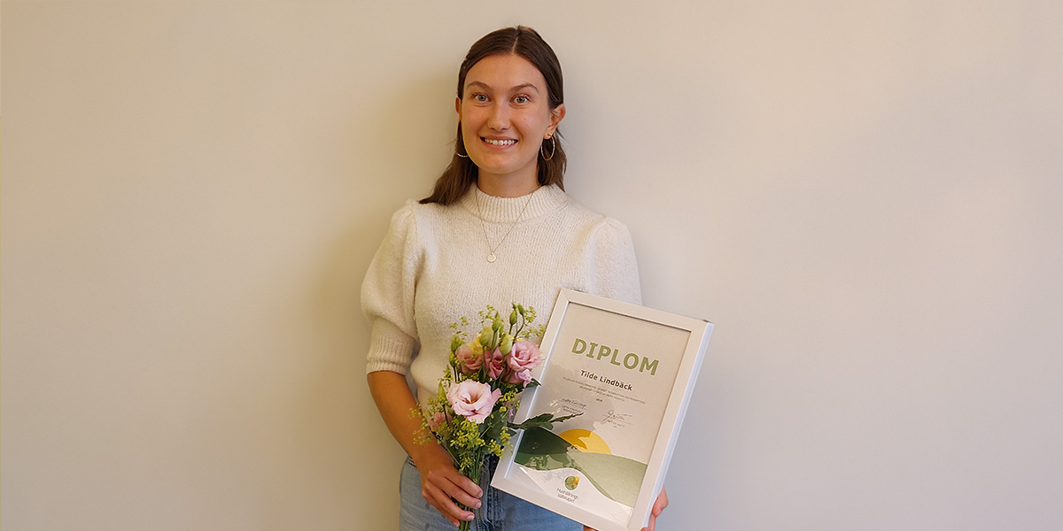 Leende kvinna står mot en ljus vägg och håller en blombukett samt ett inramat diplom. Text på diplomet: “DIPLOM” och “Tilde Lindbäck”. Porträttliknande bild som signalerar utmärkelse eller avslutning.
