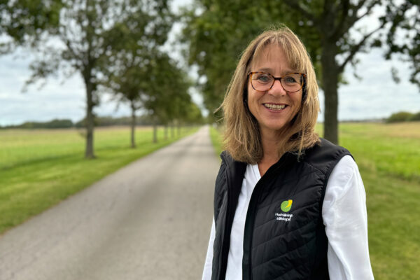 En kvinna i vit skjorta och svart väst men logotyp står i en alle och ser glad ut. Hon har brunt axellångt hår och glasögon.