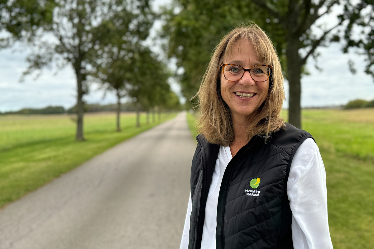 En kvinna i vit skjorta och svart väst men logotyp står i en alle och ser glad ut. Hon har brunt axellångt hår och glasögon.