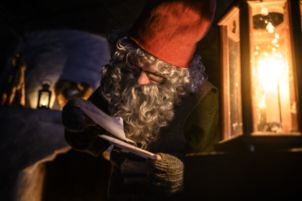 Tomtelik figur med röd luva, glasögon och lockigt grått skägg läser ett brev i händerna. Han står utomhus i mörker, upplyst av en lysande fotogenlykta till höger och snö i bakgrunden.