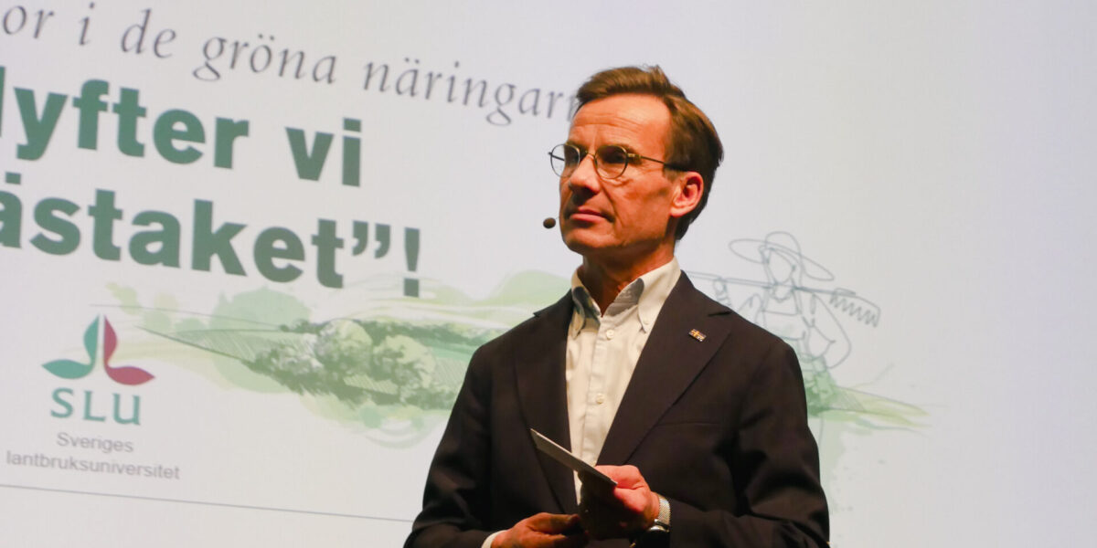 Statsministern ulf Kristersson står på scen. Skärm i bakgrunden med texten "nu flyter vi grässtaket"