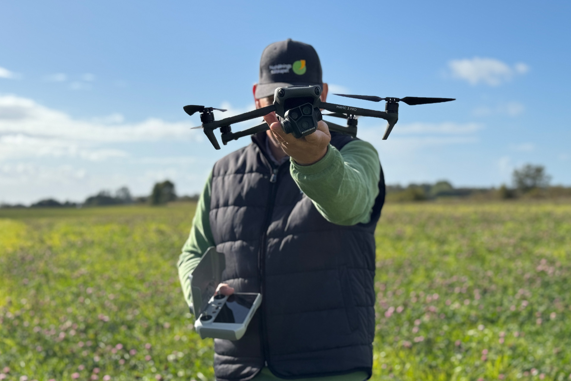 Person på en blommande åker håller fram en svart drönare mot kameran och har en fjärrkontroll i andra handen. Blå himmel och låg horisont i bakgrunden; text på drönaren: ”MAVIC 3 PRO”.