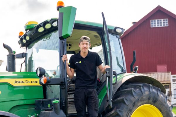 Ung man står i den öppna hytten på en grön traktor och håller i dörrkarmen, medan han tittar mot kameran. På sidan står texten “JOHN DEERE” och “155”. Röd ladugård syns i bakgrunden.