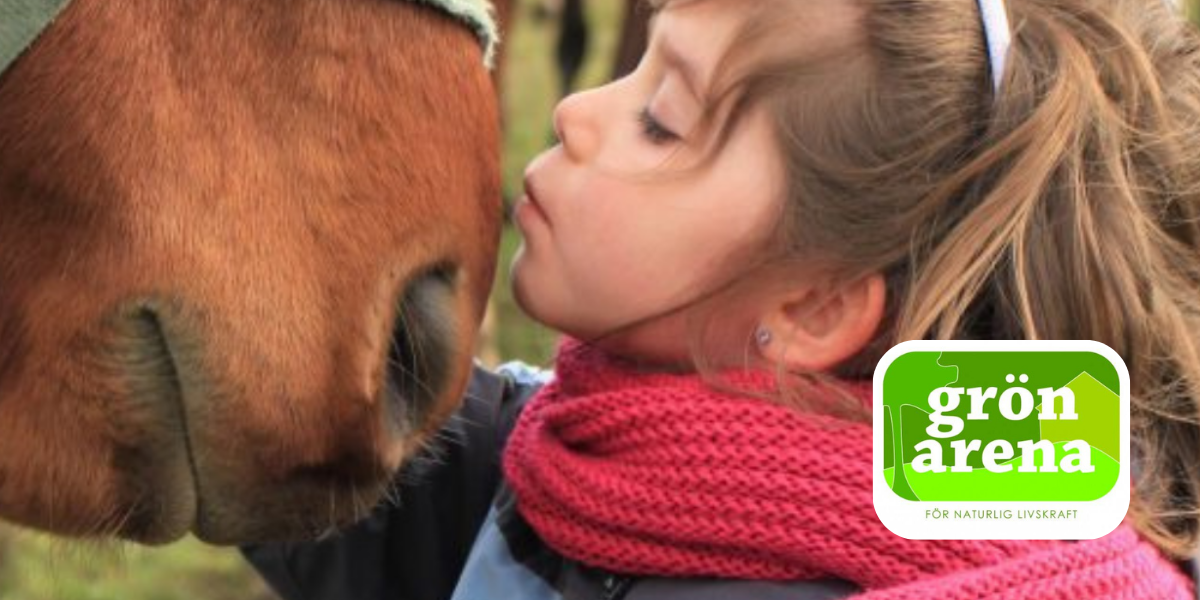 Flicka med rosa stickad halsduk och uppsatt hår lutar sig fram och pussar en hästs mule i närbild utomhus. I nedre högra hörnet syns logotypen “grön arena” och texten “FÖR NATURLIG LIVSKRAFT”.