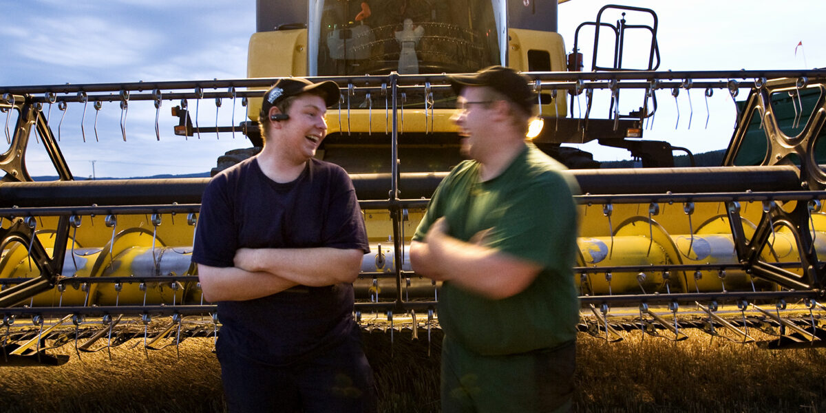 Två personer står och pratar framför en skördetröska i ett fält i skymningsljus.