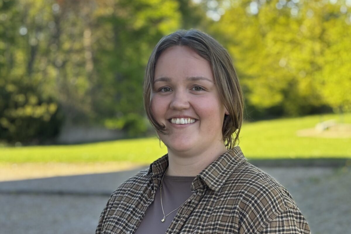 Leende person står utomhus och tittar mot kameran, klädd i brunrutig skjorta över en mörk tröja och med halsband. I bakgrunden syns en solig park med gräs och gröna träd i oskärpa.