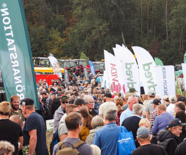 Stor folkmassa promenerar tätt mellan utställningsflaggor och jordbruksmaskiner vid en mässa utomhus. Flaggor med texten ”LÖNÅRSTATION”, ”Avida”, ”bvea” och ”SKOGSNATION” syns mot en skogsklädd bakgrund.