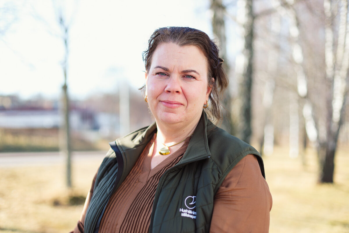 Kvinna står utomhus och ser in i kameran med ett lugnt uttryck, klädd i brun tröja och mörk väst. På västen syns texten ”Hushållningssällskapet”. Oskarp parkmiljö med träd i bakgrunden.