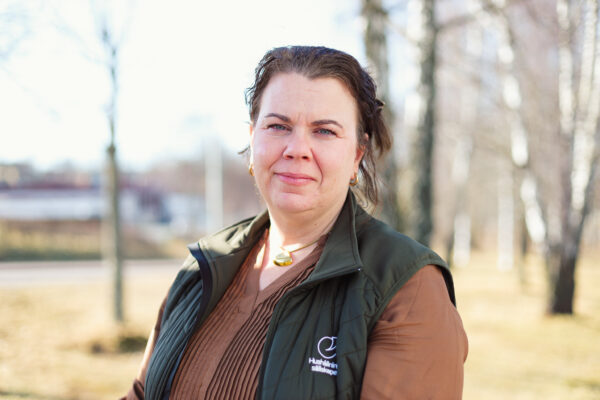 Kvinna står utomhus och ser in i kameran med ett lugnt uttryck, klädd i brun tröja och mörk väst. På västen syns texten ”Hushållningssällskapet”. Oskarp parkmiljö med träd i bakgrunden.