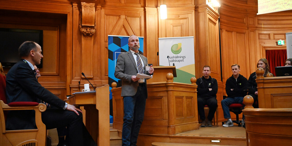 Man i kavaj står vid en talarstol och håller papper medan han föreläser i en träpanelad sal. Åhörare sitter bredvid. Bakom syns en rollup med texten ”Hushållnings sällskapet”.