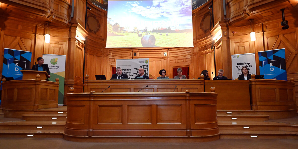 Politikerpanel sitter vid ett långt bord på en scen i en träpanelad sal, medan en talare står vid en talarstol. Ovanför visas en bild av fält med höbalar; skyltar: ”Kunskap för landets framtid” och ”KD”.