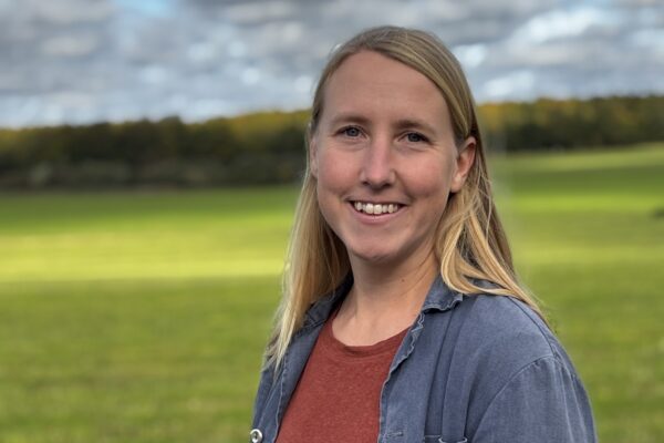 Leende kvinna med långt blont hår står vänd mot kameran utomhus, klädd i rostfärgad tröja och blå skjorta. I bakgrunden syns en grön äng, trädlinje och molnig himmel.