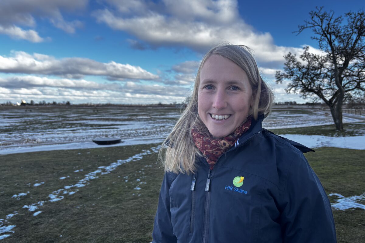 Leende kvinna i mörk jacka med loggan och texten ”HIR Skåne” står utomhus vid snötäckta åkrar, med halsduk och ljust hår i vinden. Bakom syns blå himmel med moln och ett ensamt träd.