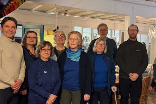 Åtta personer står samlade och poserar för ett gruppfoto inomhus, flera med glasögon och varma tröjor. De står i ett ljust rum med fönster, bord och stolar, och en ljuskrona i bakgrunden.