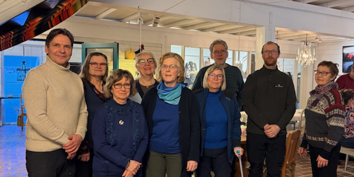 Åtta personer står samlade och poserar för ett gruppfoto inomhus, flera med glasögon och varma tröjor. De står i ett ljust rum med fönster, bord och stolar, och en ljuskrona i bakgrunden.