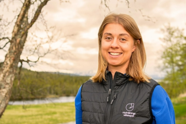 Leende kvinna står vänd mot kameran och poserar för ett porträtt utomhus, iklädd svart väst med loggan och texten ”Hushållnings sällskapet”. I bakgrunden syns träd, grön äng och en sjö under molnig himmel.