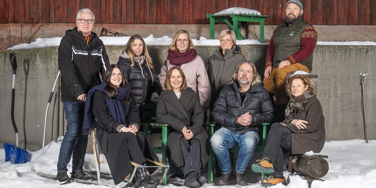 Åtta personer poserar för en gruppbild utomhus i vintermiljö, några stående och andra sittande på bänkar. De är varmt klädda och omges av snö, med en röd trävägg och gröna ställningar i bakgrunden.