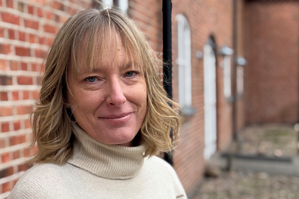 Porträtt av en leende kvinna i ljus tröja som står utomhus vid en tegelbyggnad.