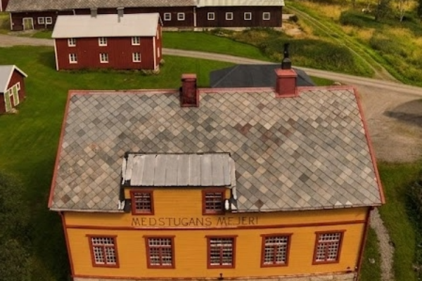 Flygvy över en gul byggnad med skylten ”MEDSTUGANS MEJERI”, omgiven av grusplan och grönska. I bakgrunden syns flera röda gårdsbyggnader och en väg som slingrar genom ett kuperat, skogsklätt landskap.