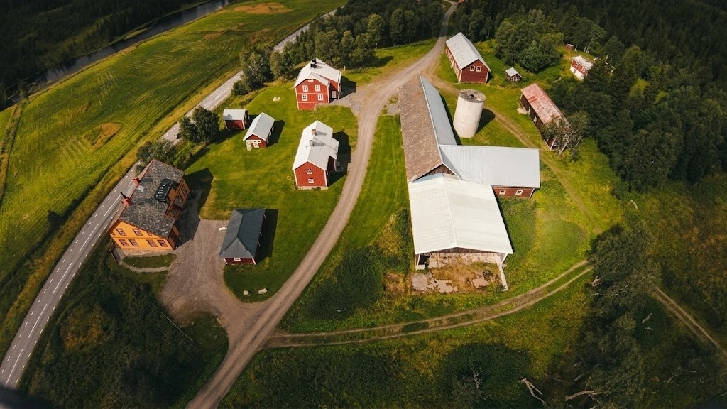 Flygvy över en lantgård med flera röda byggnader och stora vita ladugårdar, som ligger längs en grusväg. Fält och skog omger området, med en väg och vattendrag synliga i bakgrunden.