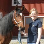 En leende kvinna står bredvid en brun häst och håller i ett blått grimskaft. Hästen har mörk man och en liten vit fläck i pannan, framför en röd stallbyggnad i solljus.