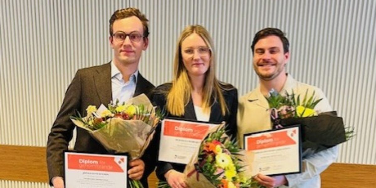 Tre personer står bredvid varandra och håller diplom och blomsterbuketter. Två män och en kvinna ler mot kameran i en inomhusmiljö med ribbad vägg. Text på diplomen: “Diplom”.