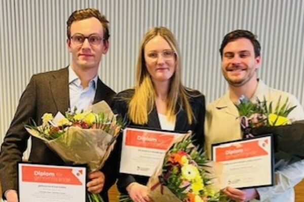 Tre personer står bredvid varandra och håller diplom och blomsterbuketter. Två män och en kvinna ler mot kameran i en inomhusmiljö med ribbad vägg. Text på diplomen: “Diplom”.