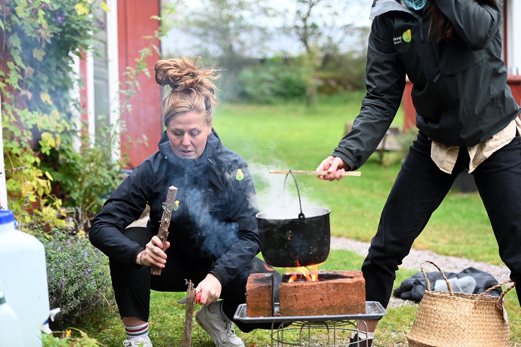 Kvinna på huk klyver ved och matar en liten eld, medan en annan person håller en svart gryta över lågorna med en pinne. Rök stiger upp; de står utomhus vid ett rött hus med gräsmatta.