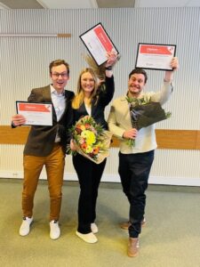 Tre glada personer står i en sal och firar genom att hålla upp diplom med texten ”Diplom”. Kvinnan i mitten håller en blomsterbukett, och mannen till höger håller en bukett, som vid en avslutning eller prisutdelning.