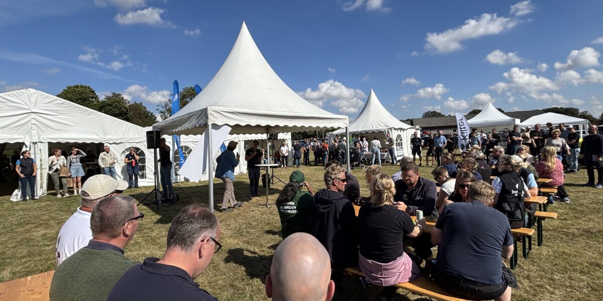 Publik sitter vid träbänkar och lyssnar på en talare vid ett mikrofonställ under ett vitt tält. Flera vita tält och stående besökare syns på en gräsplan under blå himmel med moln.