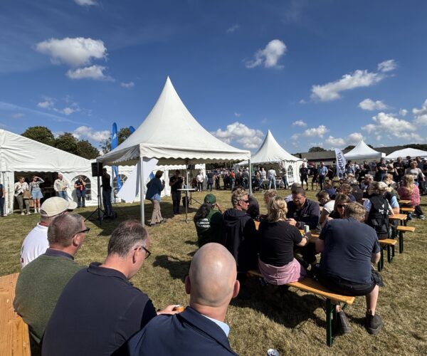 Publik sitter vid träbänkar och lyssnar på en talare vid ett mikrofonställ under ett vitt tält. Flera vita tält och stående besökare syns på en gräsplan under blå himmel med moln.