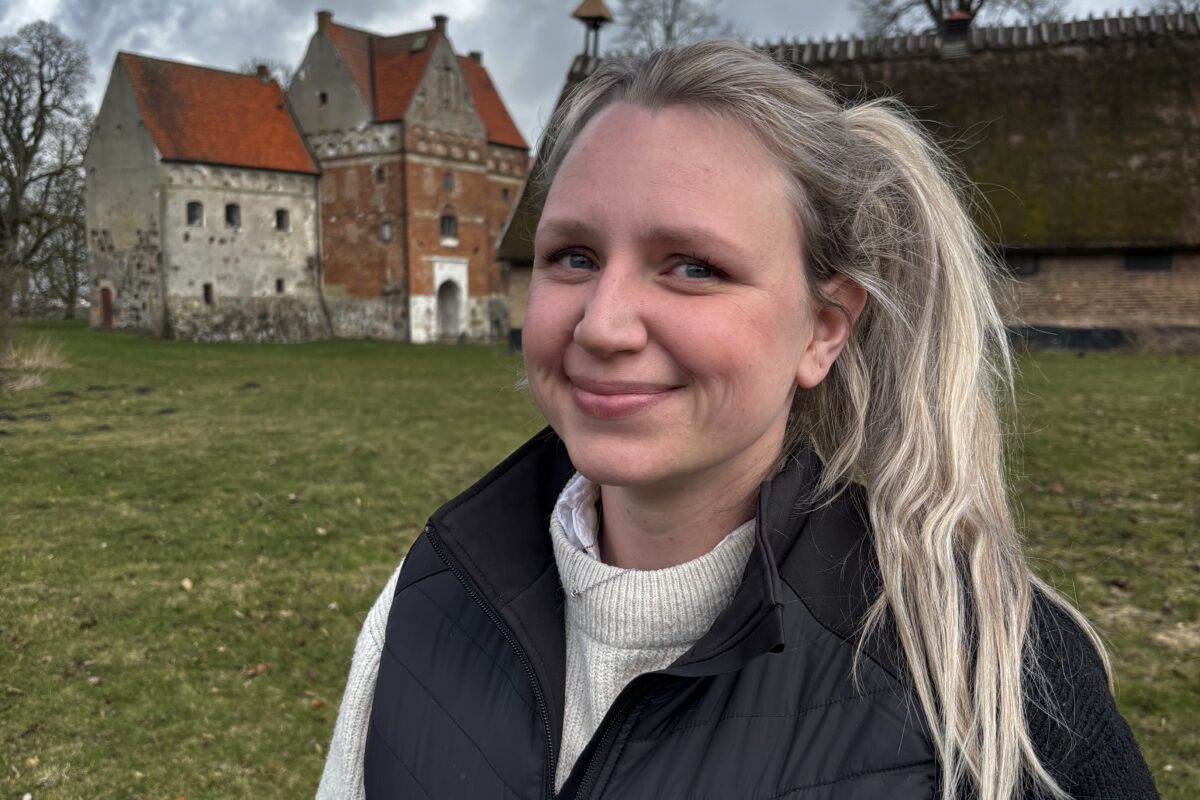 Leende kvinna står utomhus på en gräsmatta och tittar mot kameran, klädd i svart väst med logga och text “Hushållnings Sällskapet”. I bakgrunden syns en äldre borgbyggnad med röda tak under molnig himmel.