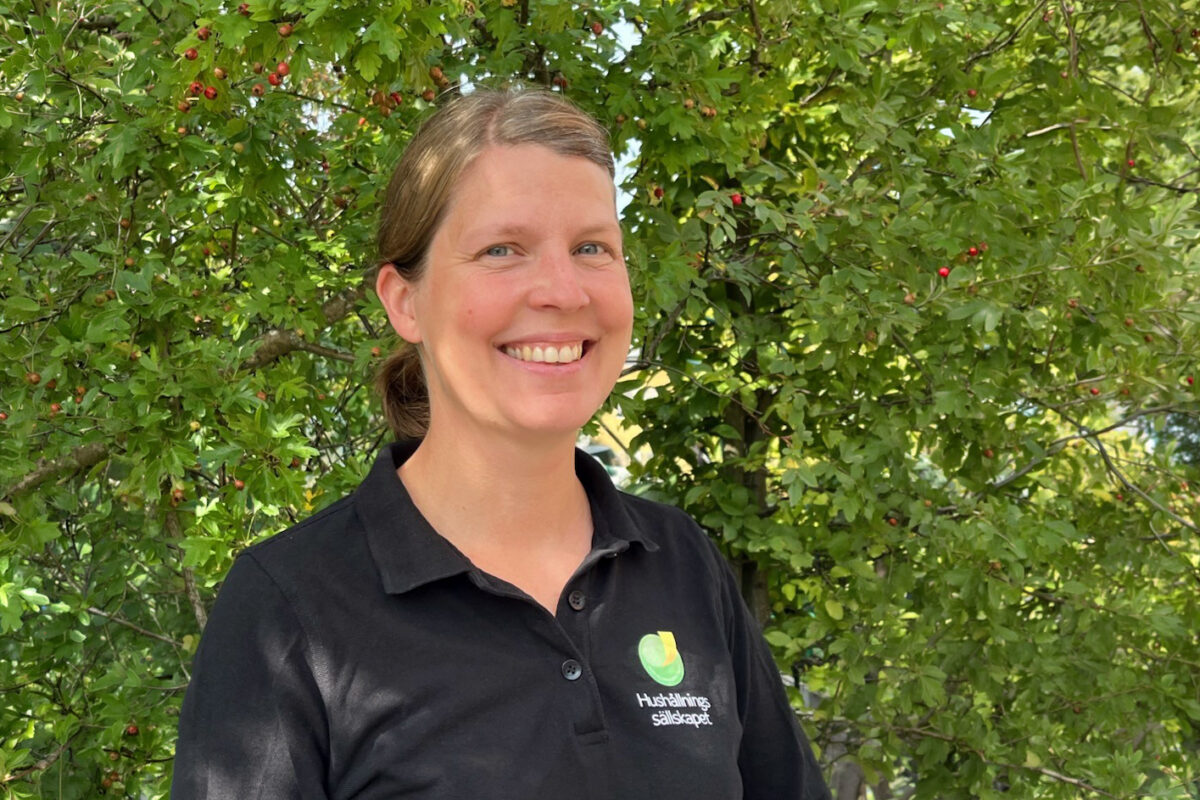 Leende person i svart pikétröja med logotypen och texten ”Hushållningssällskapet” står utomhus och tittar mot kameran, framför ett frodigt träd med gröna blad och små röda bär i dagsljus.