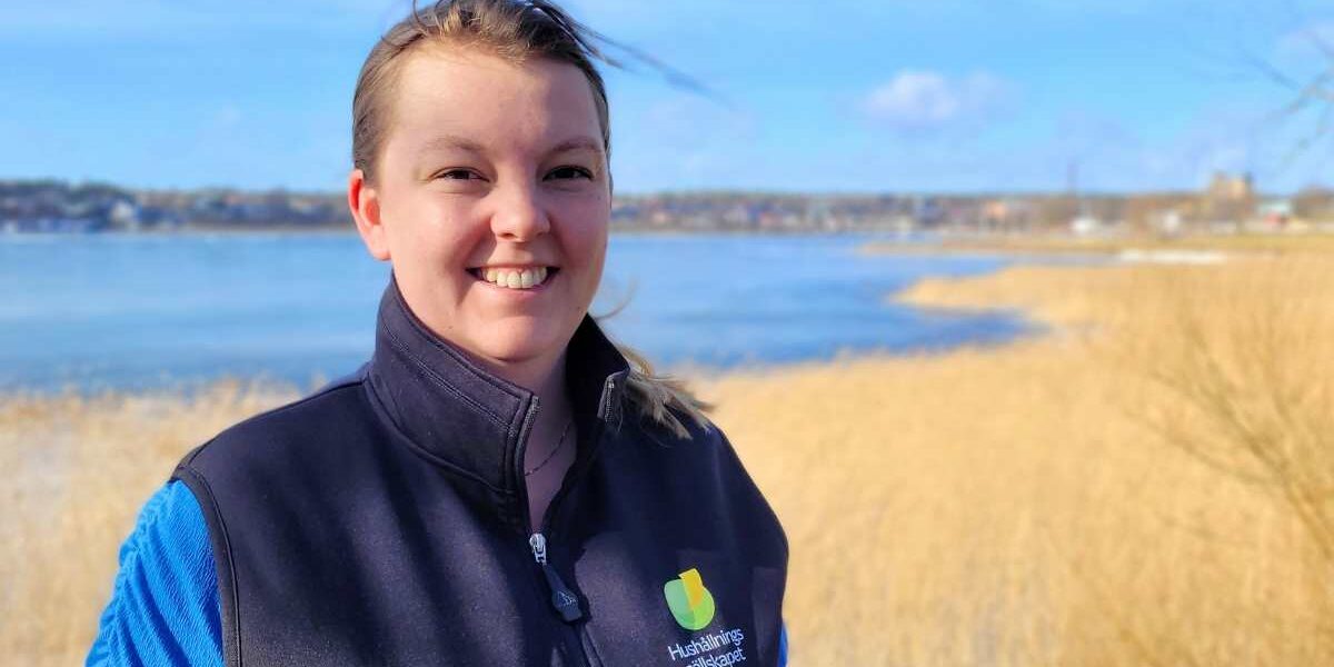 Leende person står utomhus vid en sjö och tittar mot kameran, med vinden i håret. Hen bär mörk väst med logotypen och texten “Hushållningssällskapet”; i bakgrunden syns vass, strand och bebyggelse på andra sidan vattnet.