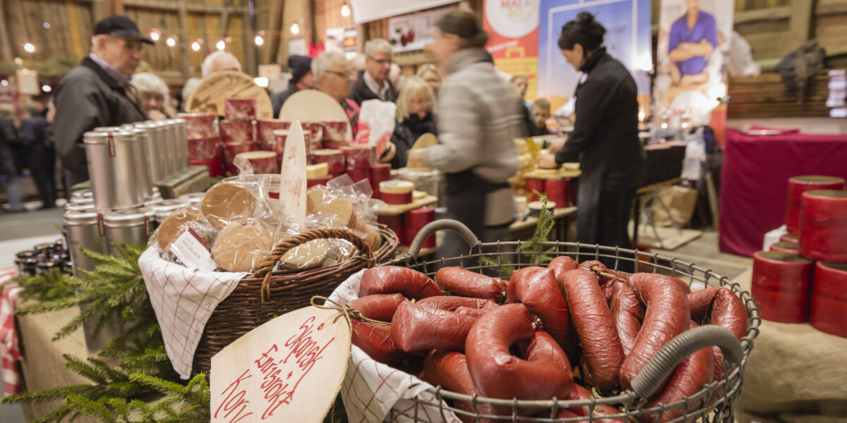 Korg med rökt korv och korg med inplastade brödkakor står på ett marknadsbord, med skyltar. Besökare handlar och samtalar i bakgrunden bland burkar och varor i en inomhusmarknad. Text: “Rökt fårkorv/älgkorv 100%”.