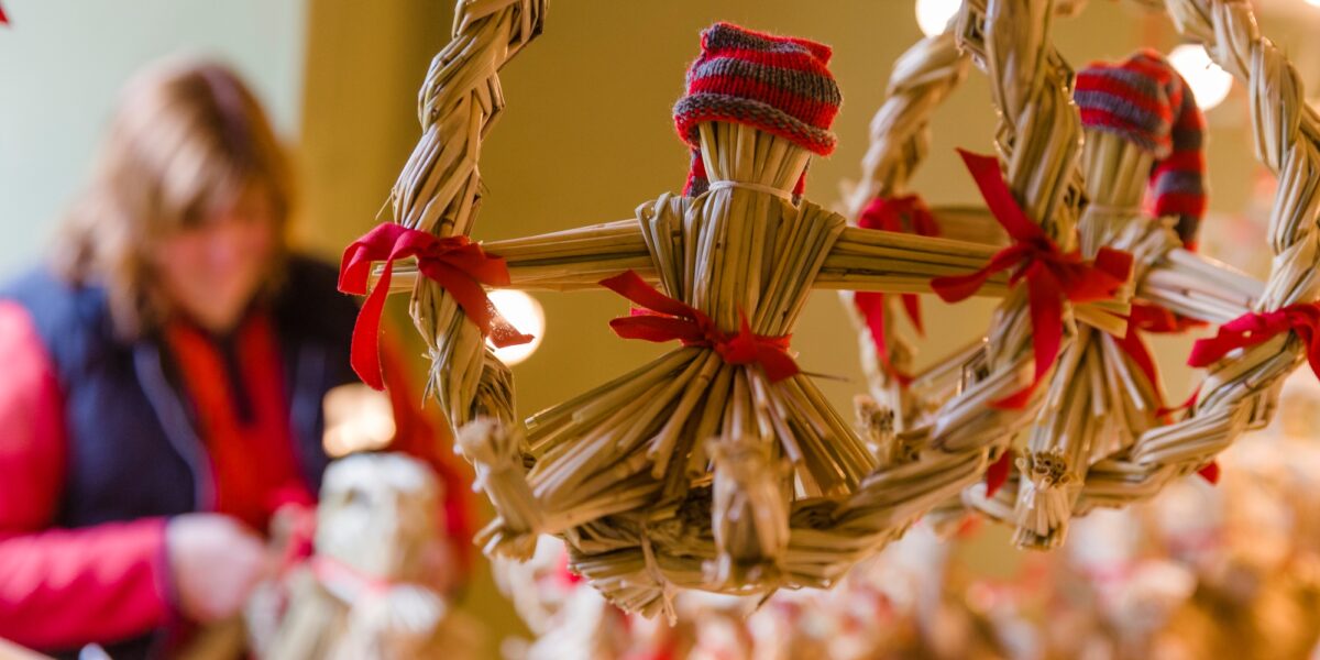 Halmbock dekorerad med röda rosetter och liten rödgrå stickad mössa hänger i en halmkrans. I bakgrunden syns en suddig person som verkar pyssla med liknande halmfigurer i varm belysning.