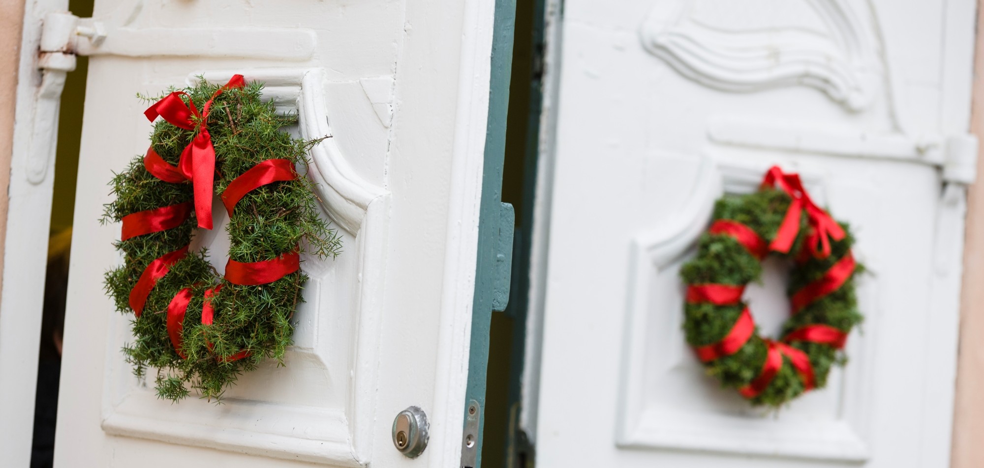 Grön julkrans med röda band sitter på en vit dörr som står på glänt. I bakgrunden syns en andra vit dörr med liknande krans, suddigt, vilket skapar en festlig entréscen.