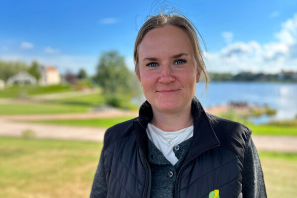 Kvinna med uppsatt ljust hår står utomhus och tittar in i kameran med ett svagt leende. Hon bär mörk väst över tröja; bakom syns grön park, gångväg och en sjö under blå himmel.