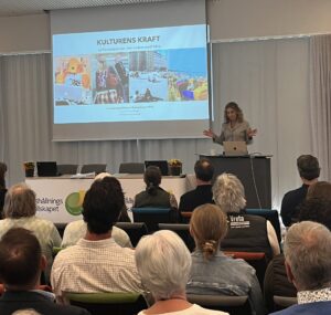 Föreläsare står vid en talarstol och gestikulerar framför en publik i en konferenssal. På projektorduken syns presentationstiteln: “KULTURENS KRAFT – varför beteenden inte ändras med fakta”, med foton och namn längst ned.