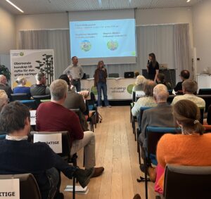 Tre paneldeltagare står och talar inför en sittande publik i en konferenssal. På projektorduken står: “Paneldiskussion – Företags förändringsresa i den gröna näringen” med “Magdalena Hermelin, Hermelins Grönsaker” och “Magnus Nilsson, Helleberga Lantbruks AB”.