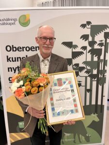 Äldre man med glasögon ler och håller en bukett blommor samt ett inramat diplom. Bakom syns loggan och texten ”Hushållningssällskapet” samt diplomet ”Östergötlands Läns Hushållningssällskap” och ”DIPLOM”, framför en illustrerad skogsmiljö.