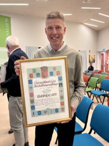 Leende man håller upp ett inramat diplom i en mötessal med stolar. Text på diplomet: ”Östergötlands Läns Hushållningssällskap”, ”DIPLOM”, ”Karl Blomqvist, Alvestad”. I bakgrunden syns andra personer och en skylt ”SLÄTTEN”.