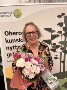 Leende kvinna med glasögon håller en stor bukett rosa och vita blommor samt ett inslaget paket. Hon står framför en rollup med texten ”Hushållningssällskapet” och ”Obero… kunsk… nytta…”, och en stiliserad skogsgrafik.