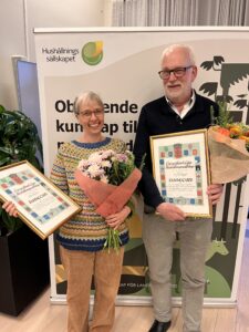 Två leende personer står framför en rollup med texten ”Hushållningssällskapet” och håller buketter samt inramade diplom med rubriken ”Östergötlands Läns Hushållningssällskap” och ”DIPLOM”, som vid en prisutdelning.