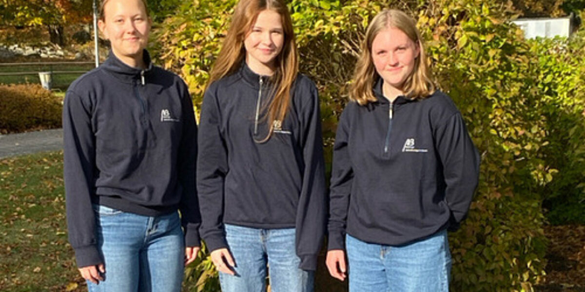 Tre unga kvinnor står bredvid varandra och ler mot kameran, klädda i mörkblå tröjor med liten logotyp och blå jeans. De fotograferas utomhus i höstsol, framför en buske och träd med gulgröna löv.