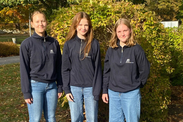 Tre unga kvinnor står bredvid varandra och ler mot kameran, klädda i mörkblå tröjor med liten logotyp och blå jeans. De fotograferas utomhus i höstsol, framför en buske och träd med gulgröna löv.