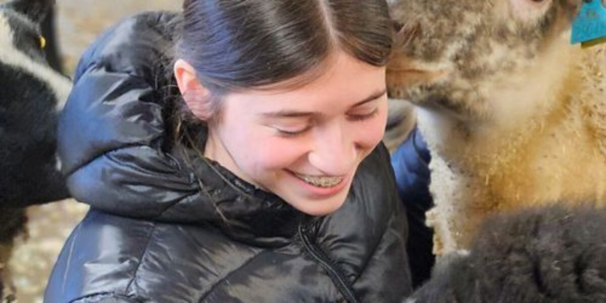 Leende ung kvinna i svart dunjacka klappar ett lamm med gul öronbricka medan ett annat lamm nosar vid hennes kind. Scenen utspelar sig i en fårhage eller ladugård med halm i bakgrunden.